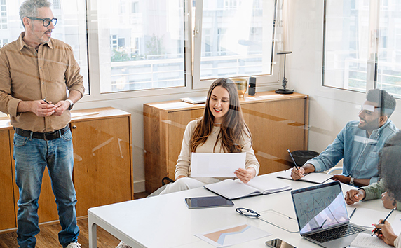 Vier Personen in einem hellen B&uuml;ro, eine steht vor einem Whiteboard, drei sitzen an einem Tisch mit Laptops und Papieren