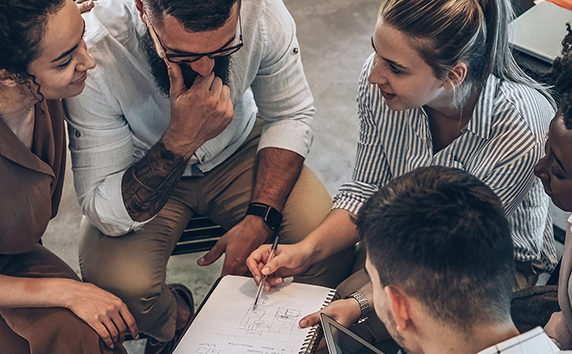 Gruppe von f&uuml;nf Personen sitzt im Kreis und diskutiert und halten ein Notizbuch in der Hand.